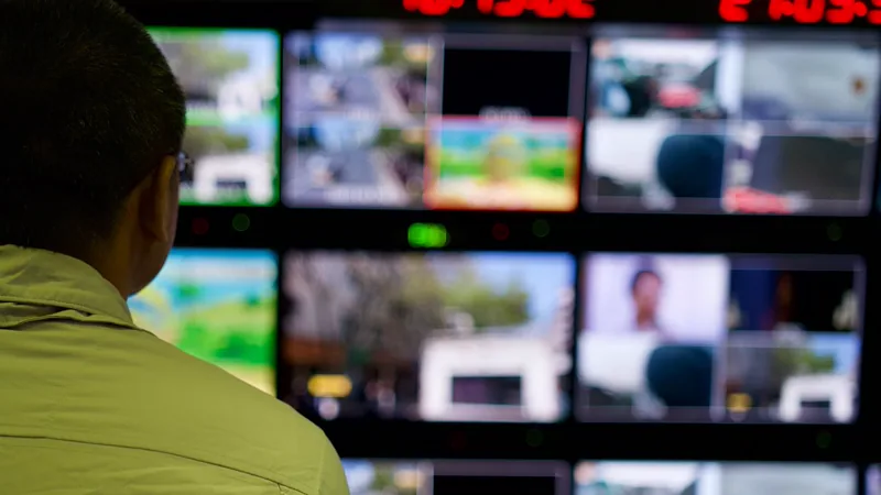 Operador de costas diante de uma parede de monitores em sala de controle, com relógios digitais no alto